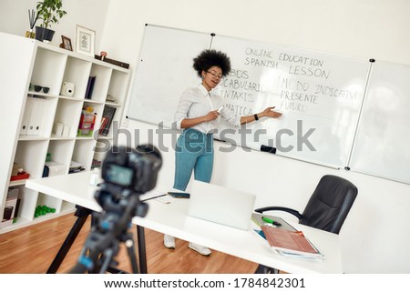 Spanish online course. Young afro american woman, female teacher standing near whiteboard teaching Spanish language online. Focus on woman. E-learning. Distance education. Recording online lesson