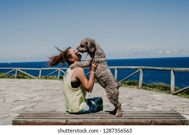 
Spanish Water Dog Licking Girl's Face In An Outdoor Park On A Sunny And Windy Summer Morning Overlooking The Sea. Lifestyle.