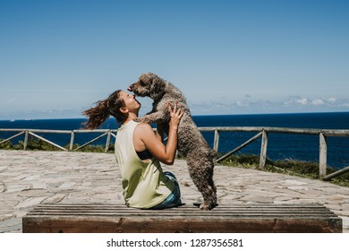 Spanish Water Dog Licking Girl's Face In An Outdoor Park On A Sunny And Windy Summer Morning Overlooking The Sea. Lifestyle.