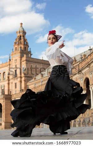 Spanish woman dancing flamenco dance in a beautiful monumental place. She is wearing a traditional black dress, a white shirt, a red flower and a headpiece.