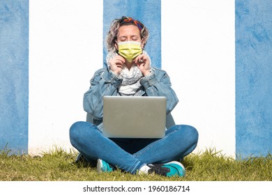 A Spanish Woman In A Face Mask Using Her Laptop Against A Blue And White Wall In The Park