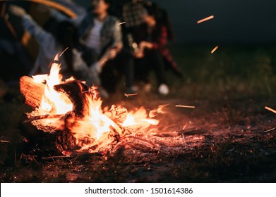 Sparking bonfire with tourist people sit around bright bonfire near camping tent in forest in summer night background. Group of student at outdoor fire fuel. Travel  activity and long vacation weekend