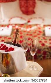 Sparking Wine, Champagne Glasses And Rose Petals On A Table In A Romantic Spa Hotel Room. Heart Shape Made On The Bed With Rose Petals.