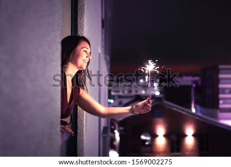 Sparkle in the night. Woman holding a sparkler out of window. New Year's Eve, Christmas party or birthday celebration at home. Happy elegant lady celebrating. City view. Light from firework stick.