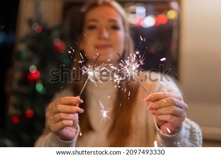 sparklers bengal fire stick in female hands holding woman in knitted wool white cozy sweater on the background of a camping trailer house outdoor on new year and christmas eve night. sparkling sparks