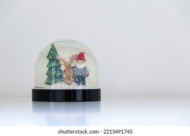 A sparkling and glittering Christmas snow globe with snow falling on Santa Claus, a reindeer, and a Christmas tree, on white snowy background, copy space on the right - Powered by Shutterstock