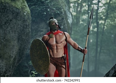 Spartan Warrior In A Battledress Walking In The Woods With His Weapon.
