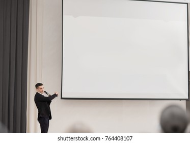 Speaker Conducts The Business Of The Conference Standing In Front Of A Large White Screen On The Stage In The Conference Room