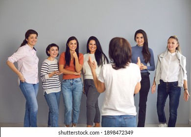 Speaker Giving Motivational Talk To Female Audience. Group Of Happy Smiling Women In Their 20s And 30s Standing And Listening To An Interesting Presentation By Professional Lifestyle Or Business Coach