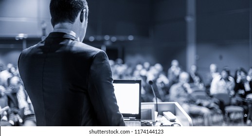 Speaker giving a talk on corporate business conference. Unrecognizable people in audience at conference hall. Business and Entrepreneurship event. Blue toned grayscale image.