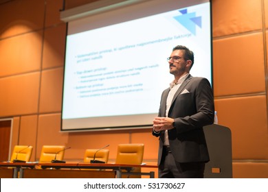 Speaker Giving Talk On Podium In Front Of White Screen At Business Conference. Entrepreneurship Club. Horizontal Composition.