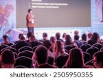 Speaker presenting to seated audience in a large conference room setting, engaging participants with insights and visuals.