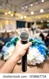 A Speaker's Hand Is Holding Wireless Microphone, Prepare For Speaking In The Education Seminar Or Business Townhall Conference On The Stage. Close-up And Mic And Hall Room As Blurred Background.