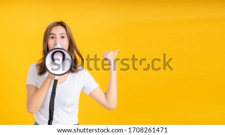 Speaking Loud noise announce of young asian woman with megaphone promotional advertising in white t-shirt smiling emotion on yellow background isolated studio shot with copy space.