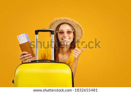 Special deal for travellers. Happy young woman in sunglasses and summer hat holding passport with plane tickets, standing next to big luggage and showing thumb up, yellow background with copy space