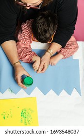 Special Education Teacher And Student In A Gown Are Painting With Their Hands A Birthday Wreath And A Yellow Cardboard With Acrylic Paints On The Table. Plastic Craft Therapy.