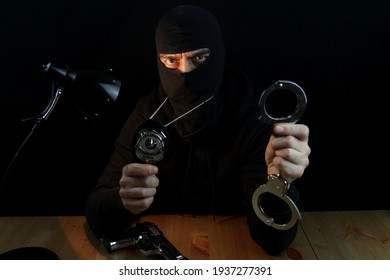 Special Forces Anti-crime Agent With Balaclava Holding Hundcuffs And Police Badge Looking At The Camera, Sitting Behind The Desk On Black Background.