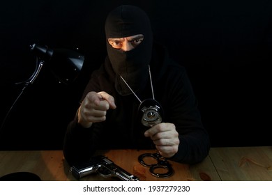 Special Forces Anti-crime Agent With Balaclava Holding Police Badge,  Pointing With Finger And Looking At The Camera, Sitting Behind The Desk On Black Background.