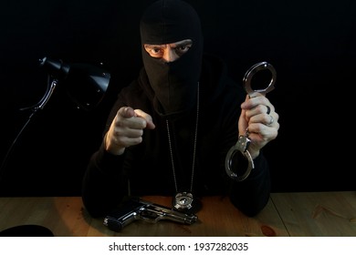 Special Forces Anti-crime Agent With Balaclava And Police Badge Holding Handcuffs,  Pointing With Finger And Looking At The Camera, Sitting Behind The Desk On Black Background.