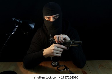 Special Forces Anti-crime Agent With Balaclava And  Police Badge Holding Gun, Looking At The Camera Sitting Behind The Desk On Black Background.