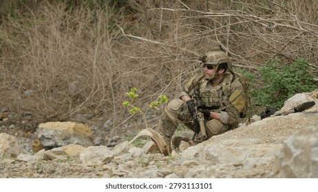 A special forces operator in full combat gear maintains a tactical overwatch position on a rugged, rocky hillside during a covert military operation.