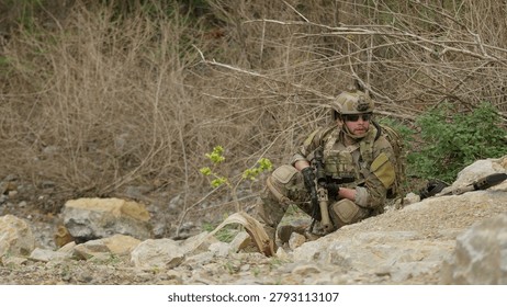 A special forces operator in full combat gear maintains a tactical overwatch position on a rugged, rocky hillside during a covert military operation.