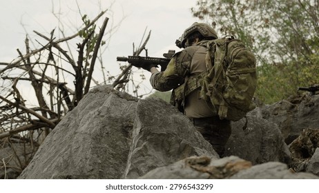 A special forces operator in full combat gear takes a defensive position, aiming an assault rifle from behind large rocks during a tactical reconnaissance mission.