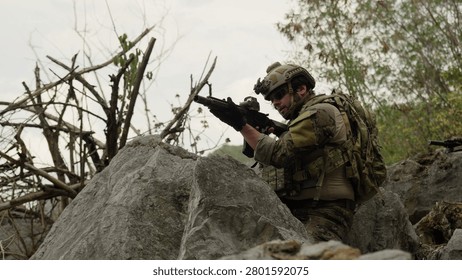 A special forces operator in full combat gear takes a defensive position, aiming an assault rifle from behind large rocks during a tactical reconnaissance mission.