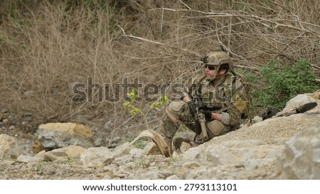 A special forces operator in full combat gear maintains a tactical overwatch position on a rugged, rocky hillside during a covert military operation.