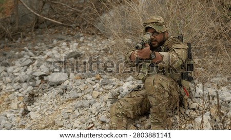 Special forces operator in full combat gear aims an assault rifle while taking cover in a rocky, desolate environment during a tactical military operation.