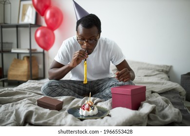 Special Occasion, Celebration And Holidays. Indoor Image Of Happy Young Dark Skinned Male Wearing Glasses And Cone Hat Having Fun In Bedroom, Celebrating His 30th Birthday, Blowing Party Horn