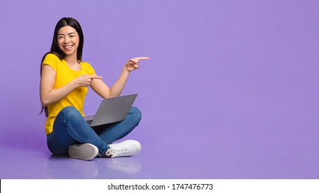 Special Offer. Cheerful Asian Girl Sitting On Floor With Laptop And Pointing At Copy Space Over Purple Background, Panorama