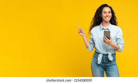 Special Offer. Portrait Of Excited Young Lady Pointing Finger Aside At Free Copy Space Holding Using Cell Phone Isolated On Yellow Orange Wall. Positive Female Showing Empty Mockup Place For Banner Ad