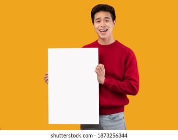 Special Offer. Portrait Of Smiling Asian Guy Holding Empty Blank Board Isolated Over Orange Studio Background. Happy Man Standing With White Vertical Square Paper Placard For Ad, Showing It To Camera