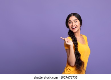 Special Offer. Portrait of smiling young indian woman with long braid pointing finger at copy space looking back isolated over purple studio background. Excited woman showing free space, place for ad