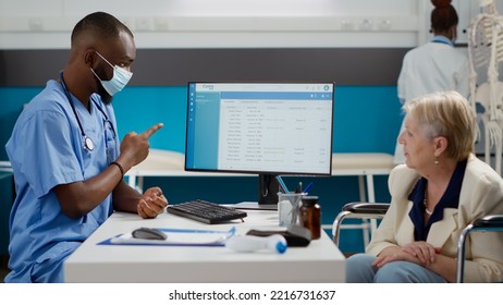 Specialist Consulting Old Woman With Disability In Office, Having Medical Appointment At Facility. Patient In Wheelchair Talking To Nurse With Face Mask About Disease And Impairment.