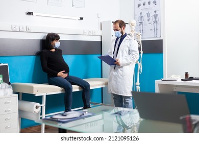 Specialist With Face Mask Consulting Pregnant Woman In Medical Cabinet. Patient Expecting Child And Doctor Meeting At Checkup Visit During Coronavirus Pandemic. Pregnancy Examination