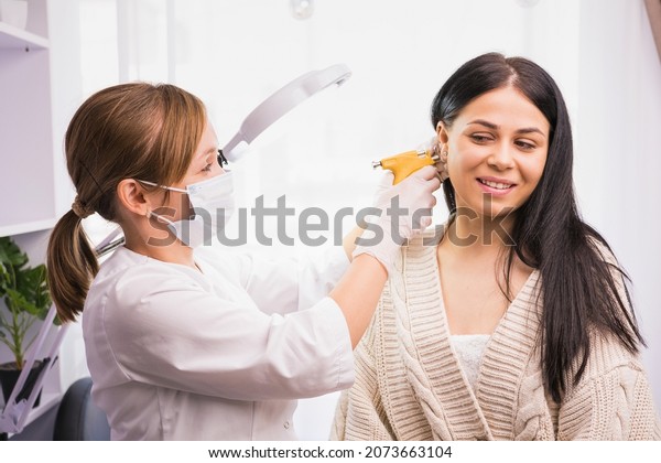 Specialist in\
medical mask and gloves pierces the client\'s ears with a piercing\
gun in the cosmetologist\
cabinet.
