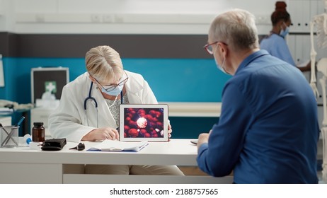 Specialist And Old Patient With Face Mask Analyzing Virus Illustration On Digital Tablet At Checkup Visit. Coronavirus Animation On Gadget To Learn About Covid 19 Prevention And Disease.