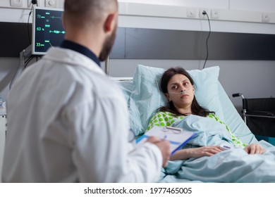 Specialist Oncologist Physician Writing Disease Examination On Clipboard Monitoring Sickness Symptom Working In Hospital Ward. Sick Patient Resting In Bed Discussing Illness Treatment