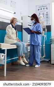 Specialist Physician Nurse Explaining Sickness Symptoms With Retired Old Woman Patient Discussing Medication Treatment During Medical Appointment In Hospital Office. Coronavirus Concept