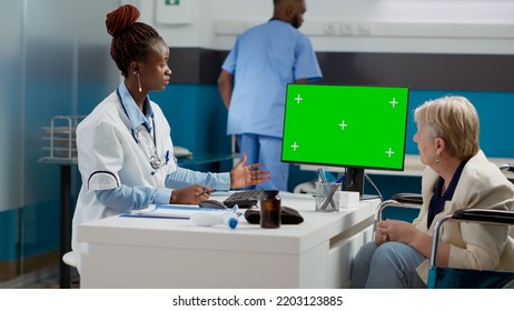 Specialist Pointing At Computer With Greenscreen Display At Checkup With Old Woman In Wheelchair. Medic And Patient Looking At Chroma Key Background With Isolated Mockup Copyspace.