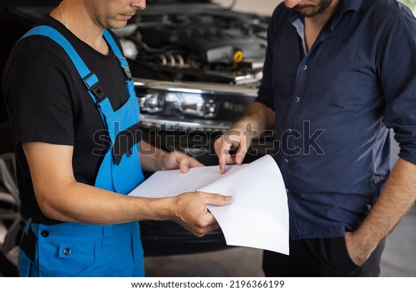 Specialist is showing info on tablet. Car\
service manager with a tablet, talking to mechanic man discussing\
car diagnostics and repairing. Mechanic talking to manager near\
vehicle in car\
service.