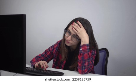 An IT specialist sits wearily in the office in front of a computer. A young girl lazily types on the keyboard. Girl with glasses and a red shirt. - Shutterstock ID 2259185759