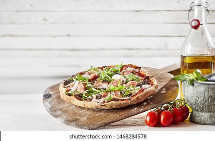 Speciality Prosciutto And Rocket Italian Pizza With Shaved Parmesan Cheese Sreved On An Old Vintage Wooden Pizzeria Paddle Alongside Fresh Ingredients Over White Wood And Copy Space