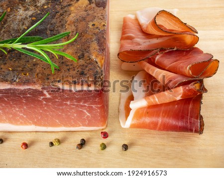 Speck with slices on the cutting board. Typical South Tyrolean smoked bacon. Sliced raw ham. Dry cured meat. Traditional cold cuts, Italian speck with rosemary and pepper.
