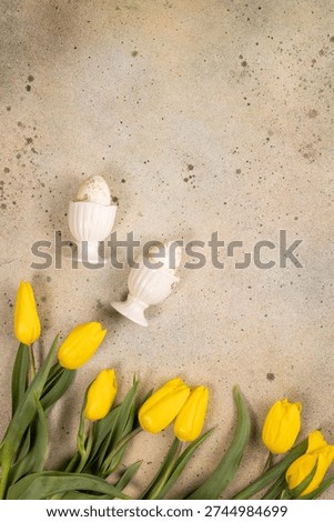 Speckled easter eggs nestled in white eggcups, surrounded by vibrant yellow tulips arranged on a rustic concrete background, creating a festive spring holiday composition with ample copy space
