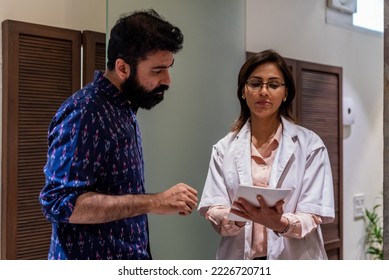 A spectacled female Indian doctor writes a prescription for a male Indian patient dressed in a casual blue shirt, while standing in the lobby hall of a medical clinic in New Delhi during the daytime. - Powered by Shutterstock