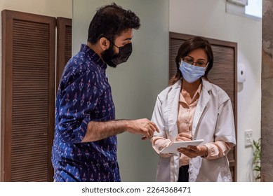 A spectacled female Indian doctor writes a prescription for a male Indian patient dressed in a casual blue shirt, while standing in the lobby hall of a medical clinic in New Delhi during the daytime. - Powered by Shutterstock