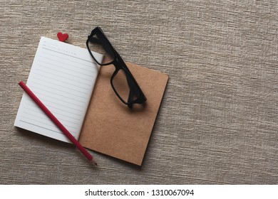 The Spectacles And Red Color Pencil On Blank Notebook, Which Lay On Velvet Bed And There Is A Red Mini Heart Book Mark